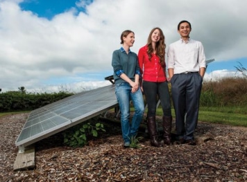 Two women and a guy standing in front of a big solar panel.