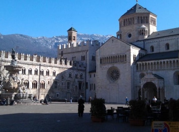 The photo depicts the Piazza Duomo in Trento, Italy. The prominent buildings visible include the Trento Cathedral (Cattedrale di San Vigilio) on the right and the Palazzo Pretorio with the Civic Tower (Torre Civica) in the background. The Fontana del Nettuno (Fountain of Neptune) is also visible on the left side of the image. The backdrop of the Alps adds to the distinctiveness of this historical and picturesque location.
