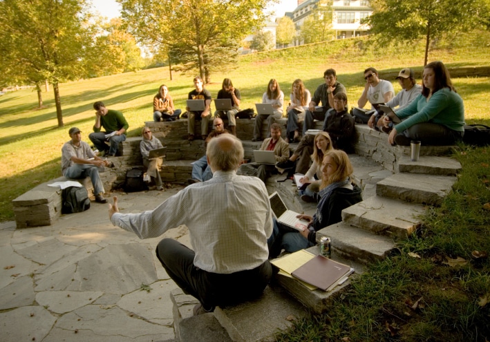 A group of students is seated in a semicircle outdoors on stone steps, attending a lecture. The setting is a grassy, tree-lined area on a sunny day.