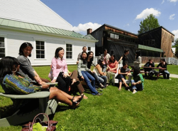 A group of people hanging out while sitting on chairs and benches outside a barn.